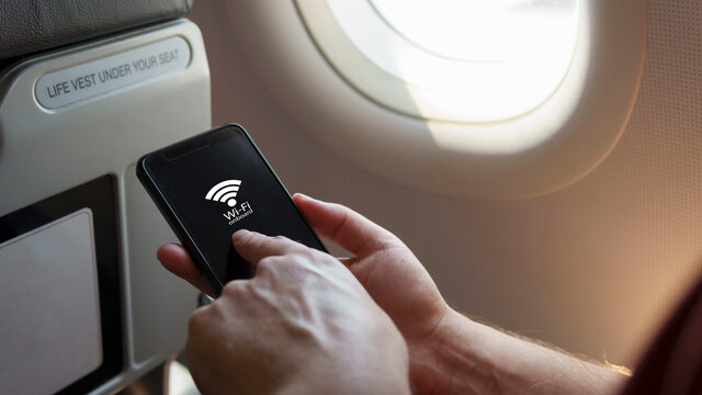 Close up Caucasian's hand holding smart phone sitting by the airplane window tap on the screen with wi-fi onboard signal. Internet wifi service during the flight, flight mode internet service concept