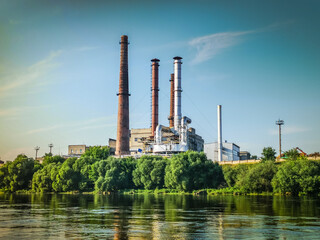 Photo of a polluting plant standing on a clean river bank among green trees in a city