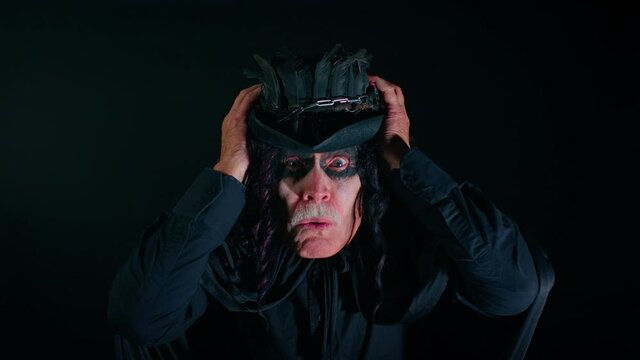 Wow. Frightening senior man with Halloween witcher makeup raising hands in surprise looking at camera with big eyes, shocked by sudden victory. Horror theme. Sinister guy on studio black background