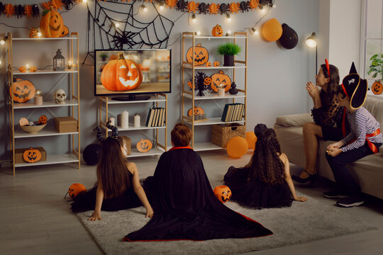 Kids in spooky costumes of witches, monsters and pirates watching children's Halloween movie sitting on floor and sofa in interior decorated with orange and black pumpkins at fancy dress party at home
