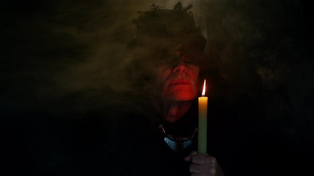 Frightening senior man with Halloween witcher makeup looking at candle, conjure, hex, wiz. Horror theme. Sinister sorcerer guy on studio black background. Voodoo magical creepy rituals with candle