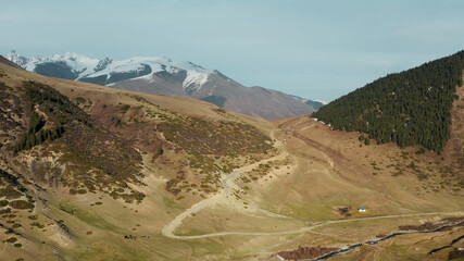 An airy picturesque landscape with a large valley with green mountains, a road and a small white house, against the background of a large mountain with snowy slopes and a blue sky with clouds. 