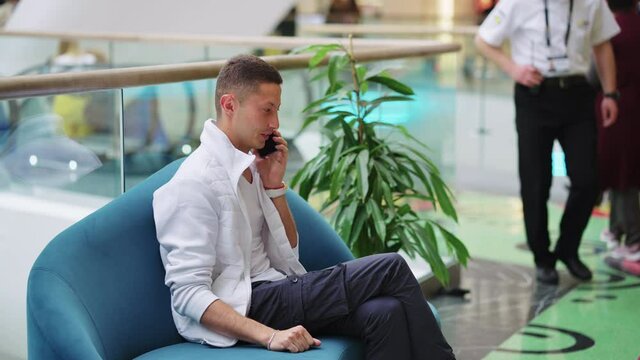 Young Man In Stylish Outfit Sitting On Sofa In Shopping Mall And Talking On Phone, Escalator With People On Blurred Background. Side View Person Using Gadget For Communication