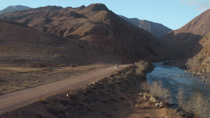 An aerial landscape of a mountain valley with a blue mountain river and a road along which a white car leaves. A picturesque landscape with large mountains against the background of a blue sky.