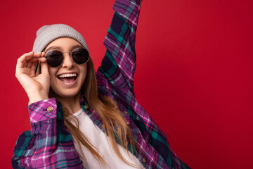 Attractive funna amazing young dark blonde woman isolated over red background wearing colourful stylish shirt grey hat and modern sunglasses looking at camera and having fun