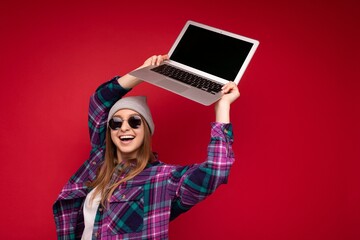 Amazing charming beautiful laughing funny smiling blond young woman holding computer laptop with empty monitor screen wearing sun glasses hat and colourful shirt looking at camera isolated over red