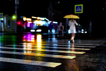 Rainy city street reflections. Unrecognizible woman with yellow umbrella cross street by crosswalk....