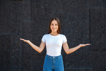 Expression, emotions, gesture and people concept - Happy Caucasian girl holding arms at her sides like a scale and standing in a white T-shirt and jeans outdoors on a black background with copy space
