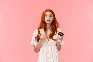 Distracted questione cute redhead girl raise head at camera to answer question, looking wondered camera, holding smartphone and credit card, trying pay for online purchase cant understand something - obrazy, fototapety, plakaty