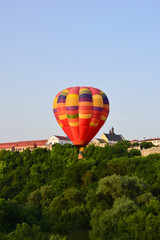 Fototapeta premium A large multi-colored balloon flies very low over the river and between dense trees. In the background, the buildings of the ancient city