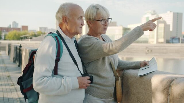 Happy old couple tourists with backpack and photo camera are looking at map then pointing at beautiful city views on summer day. Travelling and retirement concept.
