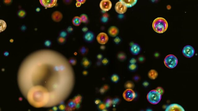 Many rainbow bright soap bubbles fly and burst on a black background. Multicolored round bubbles move in space in slow motion and shimmers in the rays of light. Close up.