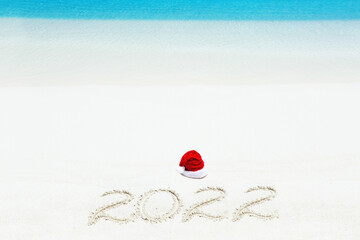 Christmas hat and 2022 inscription written on sandy beach, New Year card