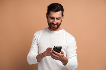 Fototapeta premium Assertive bearded man attentively looking at display of smartphone he holding, surfing internet, doing shopping online. Indoor studio shot isolated on beige background