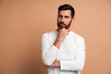 Portrait of pensive handsome bearded young man standing, touching his face, looking aside away and thinking about something. Indoor studio shot, isolated on beige background