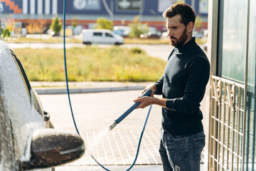 Horizontal shot of caucasian man using high pressure sprayer with cleaning foam at his black car at car wash service outdoors. Clean car outdoor concept