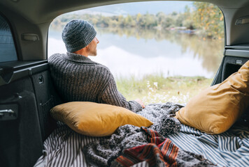 A man dressed in warm knitted clothes sitting in the cozy camper trunk and enjoying the mountain lake view. Warm early autumn auto traveling concept inside car pilows and blankets view image. - obrazy, fototapety, plakaty