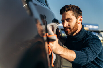 Man wiping his car at the street. Car detailing wash during the sunny day. Handsome bearded man in casual wear washing car doors and hood with microfiber cloth