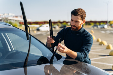 Man is changing windscreen wipers on a car while standing at the street. Male replace windshield wipers on car. Change cars wiper blades concept - obrazy, fototapety, plakaty
