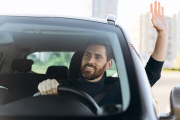 Saying hello. Handsome young smiling businessman sitting in new car and waving to someone while...