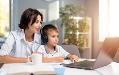 Fototapeta premium Little schoolboy in headphones sitting at desk and looking at computer screen during online lesson. Caring mother sitting near and helping her son studying at home.