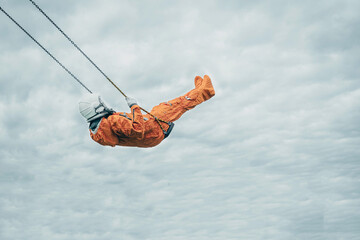 Astronaut wearing orange space suit and space helmet on a swing against cloudy sky