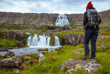 Hiker with a backpack and a red wool hat standing on a rock in front of Dynjandi waterfall in Iceland Westfjords after a successful hike. Admiring nature and powerful water flow. Iceland