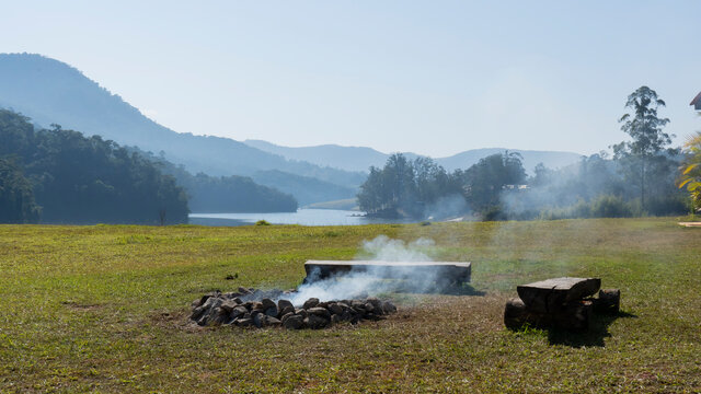 Fogueira quase apagada saindo fuma&ccedil;a em paisagem com vegeta&ccedil;&atilde;o, lago e c&eacute;u azul em perspectiva