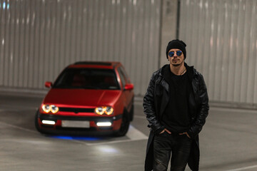 Fototapeta na wymiar Stylish brutal young handsome guy with sunglasses in fashionable black clothes with a leather jacket and a hoodie walks near a red car in a parking lot at night. Urban male casual style