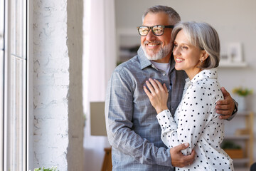 Smiling beautiful senior family couple in love embracing while standing near window at home