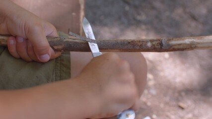 Child hand uses a penknife to remove the bark from a stick. Close up. Forest school helps children to grow in confidence as a result of the freedom, time and space they are given in their learning.  - obrazy, fototapety, plakaty
