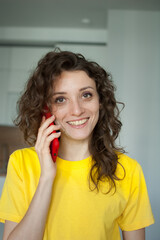 Beautiful curly girl in yellow t-shirt is talking with someone using red smartphone at home in her apartment, posing with smile, happy people concept