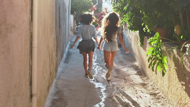 Two girls running across sunny greek street. Best friends spend summer time together. The girls are happy and enjoy life. The road by car to the sea. Friendship and love forever. The beauty of Greece.