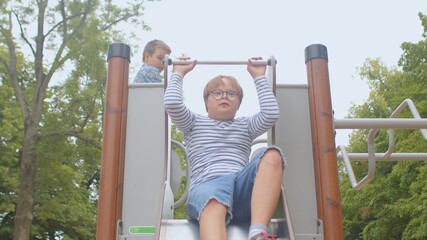 Obraz premium He looks ahead. Down syndrome boy rides on the playground slide. A bright moment of childhood joy. Dexterity of movement down.
