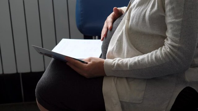 Young Pregnant Woman In Medicine Protective Mask Reading On Couch Waiting For Appointment Medical Control On Hospital. Visit Gynecologist Doctor At Clinic. Examine Expectant Belly Healthcare Check Up