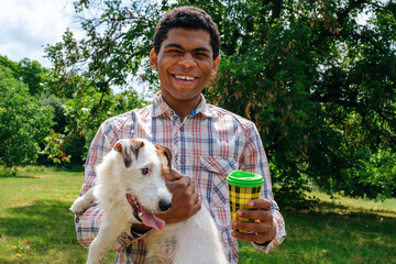 Young smiling African American man holds a Jack Russell terrier dog in his arms and drinks coffee from a disposable cup on a walk in the park, summertime. 