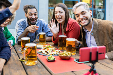 Mixed race group of young people laughing doing a video call with friends on mobile phone while drinking beer and having brunch at brewery bar - Technology and friendship concept