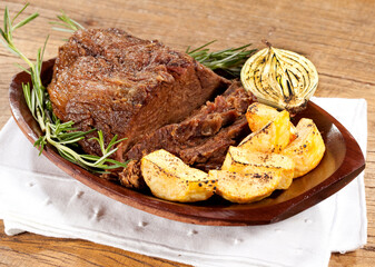 Carne assada com batatas na travessa de madeira no fundo de mesa rústica.
