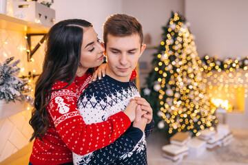A guy with a girl in a sweater and jeans hug and kiss on the background of a Christmas tree in the room