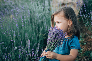 Little girl 3-4 with dark hair in denim dress sits with bouquet of lilac lavender sniffing it