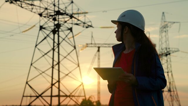 A woman power engineer in a safety helmet checks the power line using a digital tablet. Electricity company employee woman working outdoors, servicing high voltage electrical lines at sunset