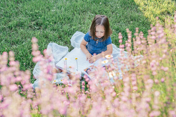 Fototapeta na wymiar Partially blurred little girl 3-4 with dark hair in denim dress sits and reads book, on lawn near flowers