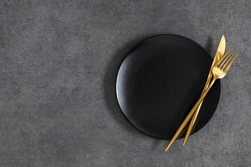 Food concept, empty black plate with gold knife and fork on gray stone table. Flat lay, top view,...