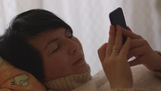 A woman uses a mobile phone in her hands while lying on the bed. Free time at home, focus of attention is the screen of the gadget. Checking online notifications.