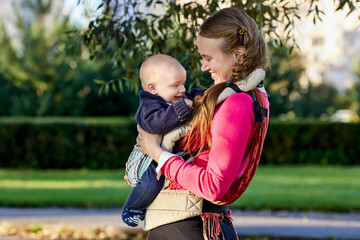 Child smiles in baby sling in hands of his mother during walking in park.
