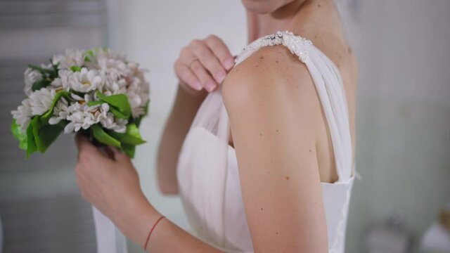 Side view unrecognizable slim young woman touching wedding dress strap smiling. Happy Caucasian bride standing with bridal bouquet of flowers indoors at home. Slow motion