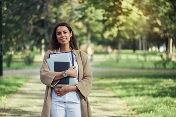 Pretty student woman is standing with the folder, notebook and laptop at the university campus...