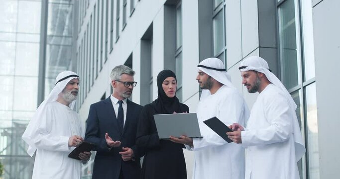 Waist up portrait of young multi ethnic entrepreneurs standing near the modern office at team meeting and speaking. Caucasian man discussing business ideas with muslim colleagues at the conference