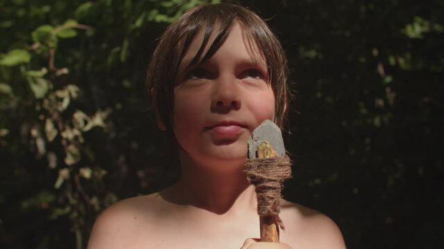 Portrait of a boy with a handmade spear in the forest. Long haired preteen playing survival game, explore, master crafting. The spot of the sun illuminates the face. The stone tip is tied with a rope