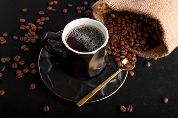 Una taza de café negro con una cucharita dorada al costado, y granos de café esparcido  